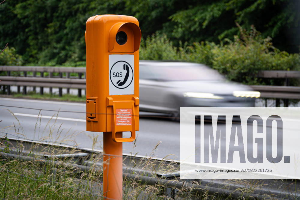 Landkreis Osnabrück, Deutschland 10. Juni 2025: Im Bild: Eine markante orangefarbene Notrufsäule steht im Vordergrund am Rand einer Straße, die vermutlich eine Autobahn oder Schnellstraße ist. Auf der Säule ist deutlich das weiße SOS -Zeichen mit einem Telefonhörer-Symbol zu sehen. Unterhalb des Hauptsymbols befindet sich eine Klappe mit der Aufschrift Notruf . Im Hintergrund rauscht ein dunkles Auto mit Bewegungsunschärfe vorbei, was die Dynamik und Geschwindigkeit des Verkehrs auf der Straße unterstreicht. Üppiges Grün auf der gegenüberliegenden Seite der Fahrbahn und Leitplanken vervollständigen die Szene. Niedersachsen