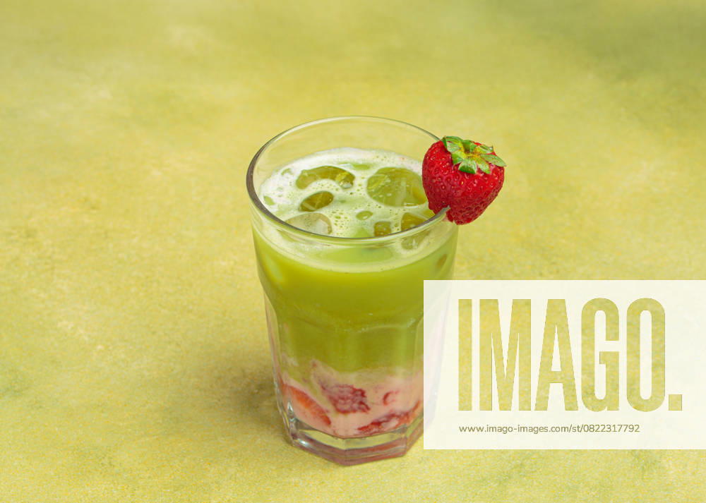 Geeister Matcha Latte mit Erdbeerpüree und Eiswürfeln, dekoriert mit frischen Erdbeeren in einem Glas auf grünem Hintergrund Refreshing iced matcha latte blended with strawberry puree and ice cubes, garnished with a fresh strawberry, served in a tall glass against a vibrant green background (License=RF)