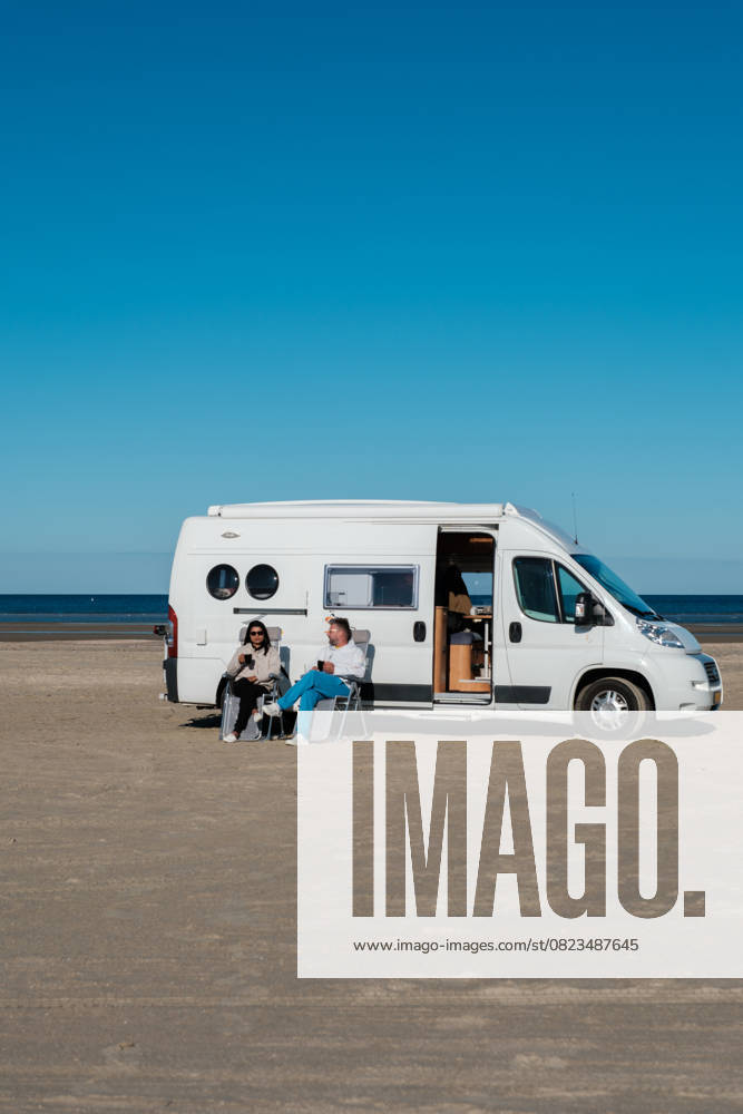 Entspannen am Strand in Dänemark mit einem Wohnmobil an einem sonnigen Tag A couple enjoys a peaceful day at a beach in Denmark, lounging outside their camper van. The clear blue sky enhances their relaxing atmosphere as they savor the tranquil surroundings. (License=RF) ,model released, Symbolfoto