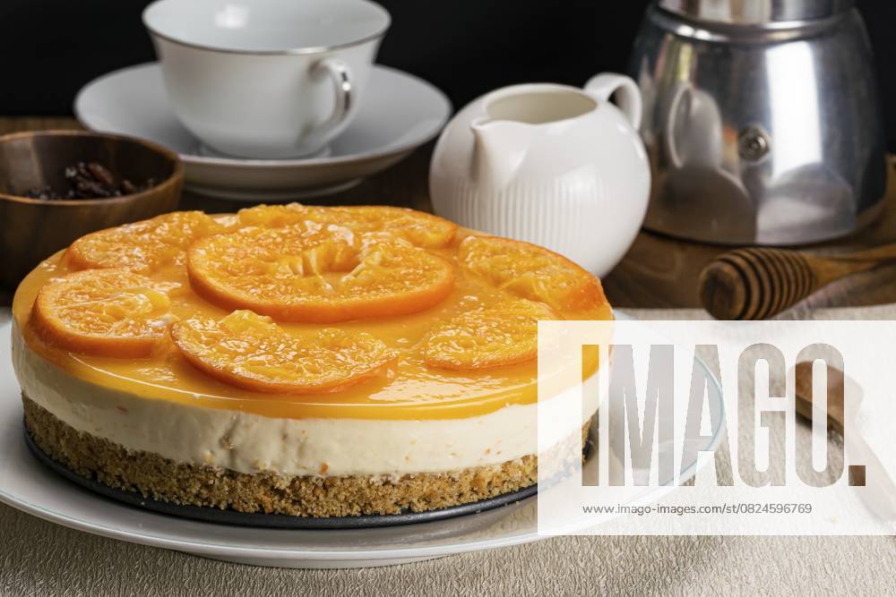 Seitenansicht eines köstlichen hausgemachten Orangenkuchens, garniert mit Stücken reifer Orangen in einer weißen Keramikschale auf einem Untersetzer mit einer hölzernen Tasse mit Rosinen, einer weißen Kaffeetasse, einem Keramik-Milchkännchen, einem Honiglöffel und einer Moka-Kaffeemaschine Side view of delicious homemade orange cheesecake garnished with pieces of ripe orange in white ceramic dish on table mat with wooden cup of raisin, white coffee cup, ceramic milk pot, honey dipper and moka pot coffee maker