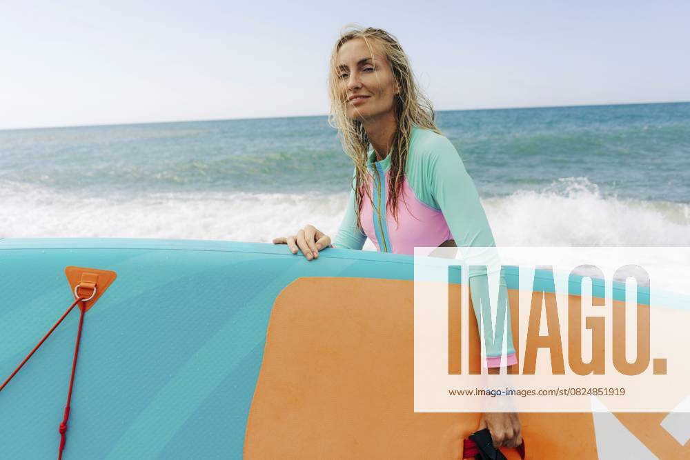 Eine junge, sportliche, blonde Frau steht am Strand und hält ein SUP Board Eine junge, sportliche, blonde Frau steht am Strand und hält ein SUP-Board