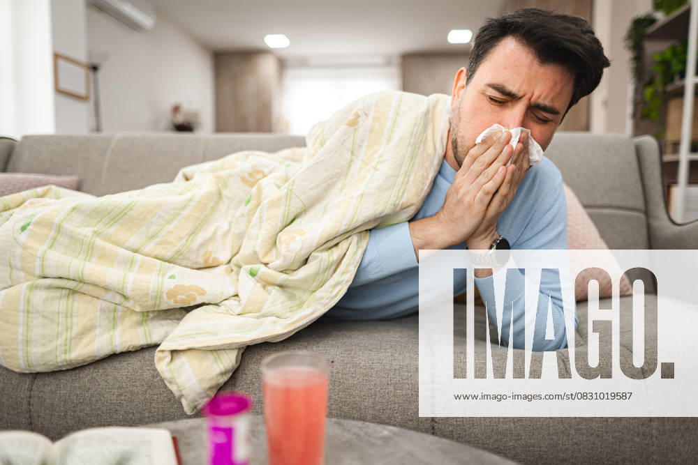 Sick man having a runny nose while lying on a sofa covered with blanket Man is seen laying on a couch, covering his face with a tissue, as he blows his nose due to sickness or allergies. His eyes may appear watery or tired., model released, , 63184974.jpg RECORD DATE NOT STATED