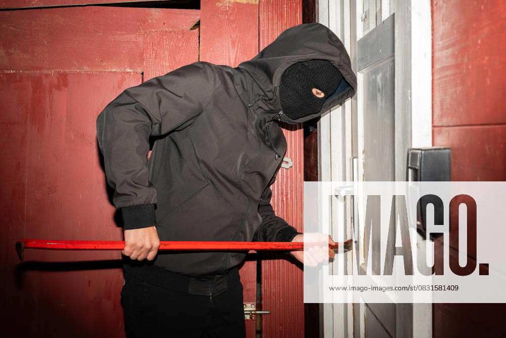 Ein vermummter Mann versucht mit einem Brecheisen eine Haustür aufzubrechen. Symbolbild die Gefahr von Einbrüchen und das Risiko für Häuser und Wohnungen durch Kriminalität