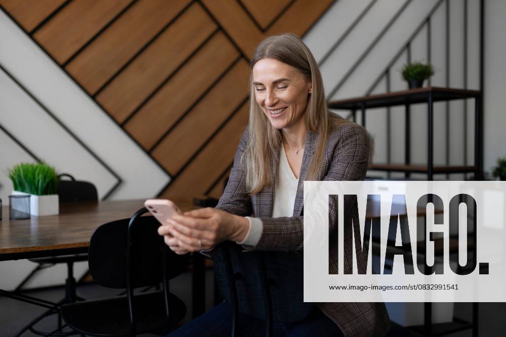 Professional woman smiling while using smartphone in modern office setting during daytime A woman with long hair and a blazer smiles while looking at her smartphone in a stylish office. Natural light highlights her relaxed demeanor among contemporary decor and furniture., model released, , 95412200.jpg RECORD DATE NOT STATED