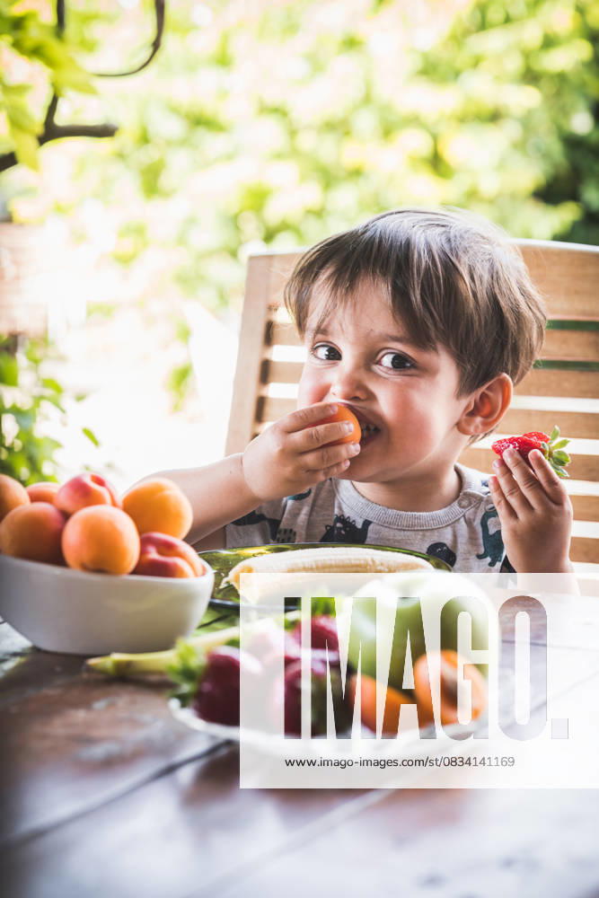 Young boy enjoying fresh fruit at a table in the garden, surrounded by vibrant greenery and various fruits. Model Released SylviePm_ID31478_1070256_001