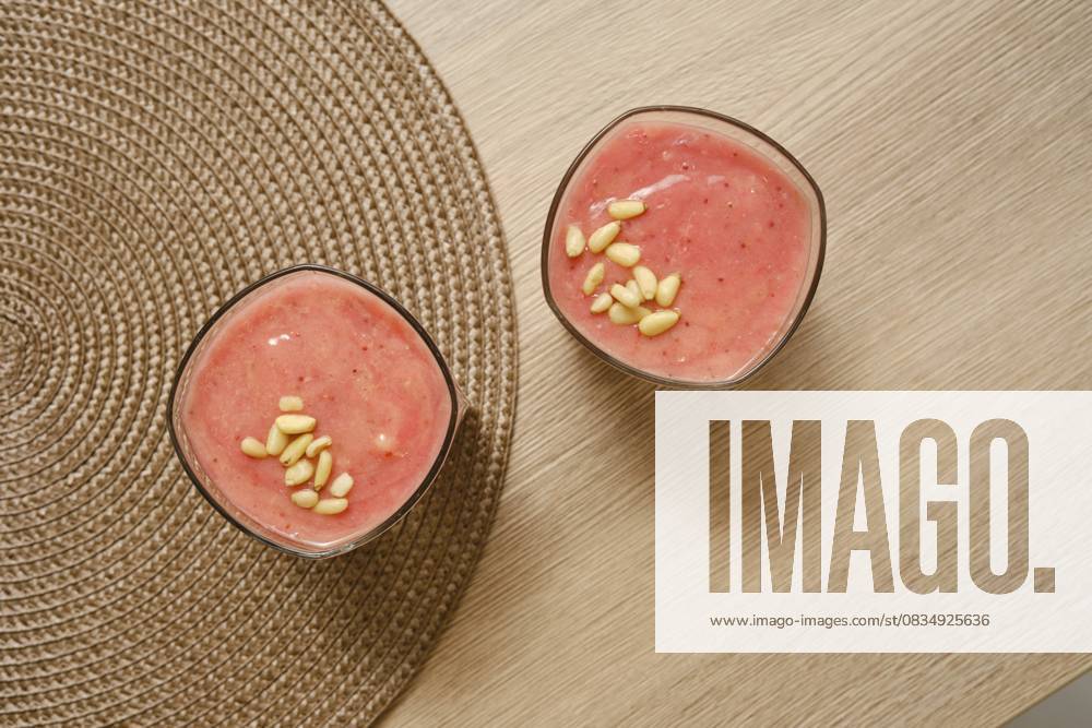 Zwei Gläser, gefüllt mit einem cremigen Bananen Erdbeer Smoothie, stehen auf einem naturfarbenen Tisch. Mit Pinienkernen garniert, ist dieses erfrischende Getränk perfekt für ein gesundes Frühstück oder einen Snack Two glasses filled with a creamy banana-strawberry smoothie sit on a natural-colored table. Topped with pine nuts, this refreshing drink is perfect for a healthy breakfast or snack