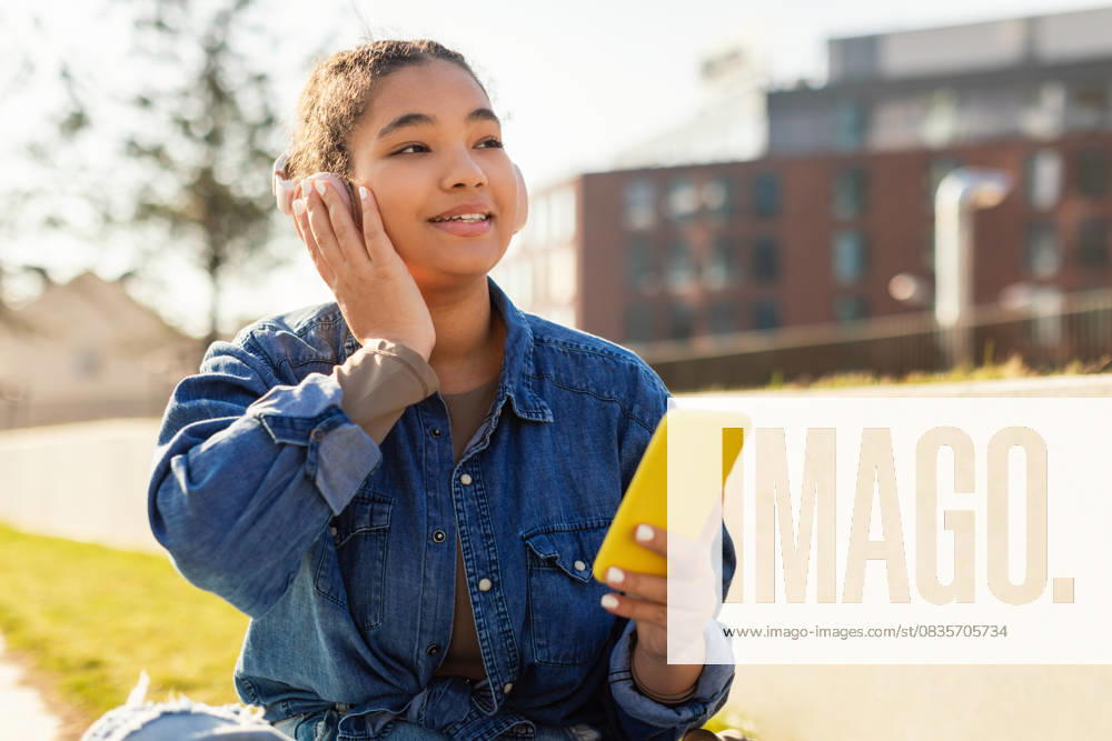 Girl Sitting on Ground Looking at Cell Phone A young girl with headphones is seated on the ground, engrossed in her cell phone screen. Her attention is solely focused on the device as she scrolls and types on the screen., model released, , 59453930.jpg RECORD DATE NOT STATED