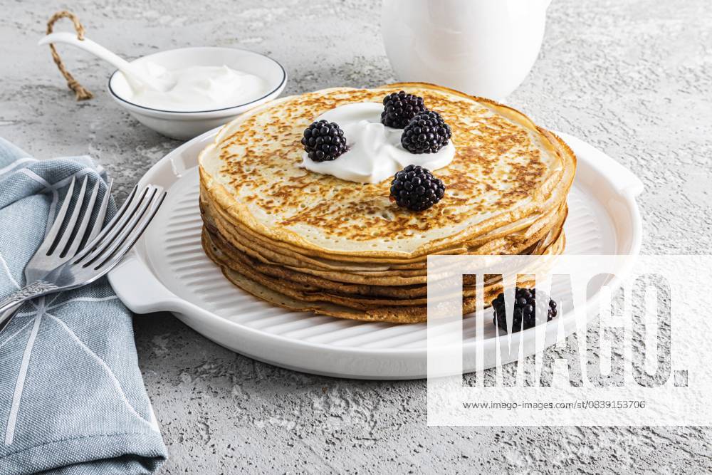 Ein großer Stapel köstlicher heißer Pfannkuchen mit saurer Sahne und Brombeeren auf einem schönen weißen Serviertablett mit Besteck und einem Grundstück. View from front A large stack of delicious hot pancakes with sour cream and blackberries on a beautiful white serving tray with cutlery and a plot. Front view