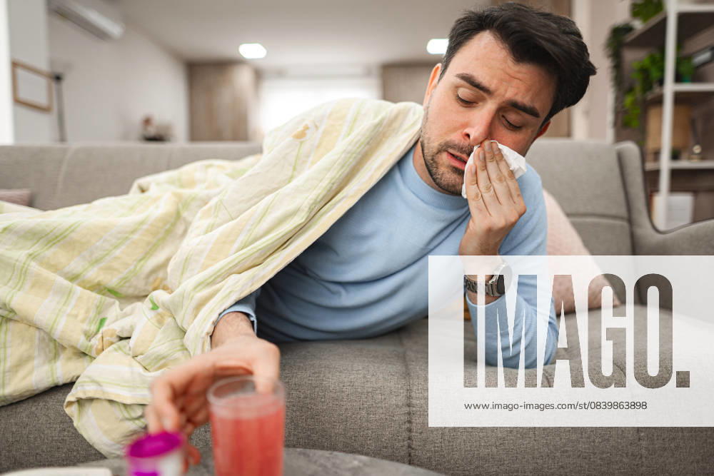 Sick man having a runny nose while lying on a sofa covered with blanket Man is seen laying on a couch, covering his face with a tissue, as he blows his nose due to sickness or allergies. His eyes may appear watery or tired., model released, , 62041996.jpg RECORD DATE NOT STATED