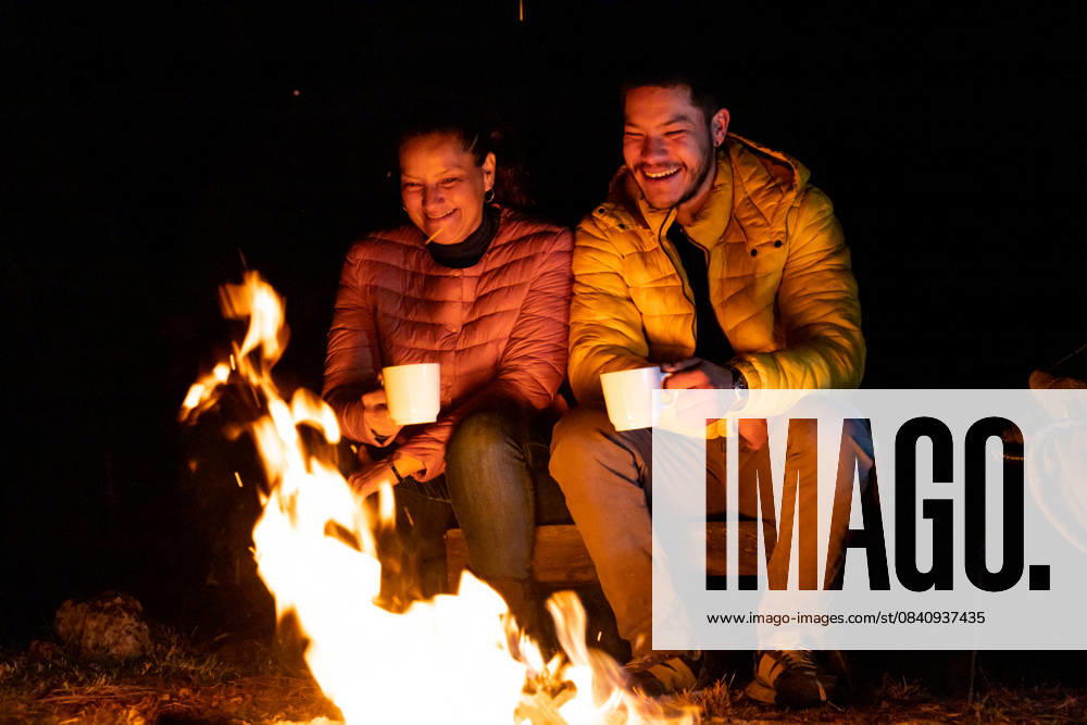 A man and a woman enjoy a cozy campfire under a starlit sky, sipping warm drinks from white mugs in the forest. The orange flames provide warmth and light in the tranquil night. Model Released EduardoGavira_ID26515_765388_029
