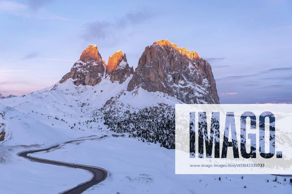 Der Langkofel im rötlichen Licht bei Sonnenaufgang. Im Vordergrund schlängelt sich die Passstraße S durch die Landschaft. Die verschneite Umgebung lässt Bergsteiger und Skifahrer vom ewigen Winter träumen. Die Dolomiten gehören zu den schönsten Skigebieten, St. Christina in Gröden, Trentino, Südtirol, Italien The Sassolungo in reddish light during sunrise. In the foreground, pass road S meanders through the landscape. The snowy surroundings make mountaineers and skiers dream of eternal winter. The Dolomites are among the most beautiful ski areas, St.Christina in Gröden, Trentino, Southtirol, Italy