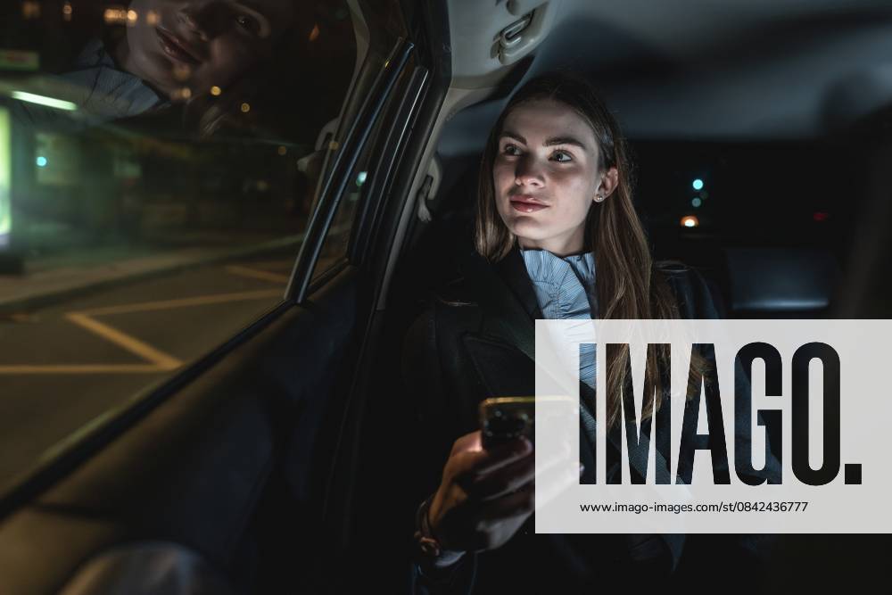 Frau, die nachts auf dem Rücksitz eines Autos unterwegs ist, ein Smartphone in der Hand hält und aus dem Fenster schaut, während sich die Lichter der Stadt auf dem Glas spiegeln und die städtischen Verkehrsdienste darstellen Woman travelling in the back seat of a car at night, holding a smartphone and looking out the window with city lights reflecting on the glass, representing urban transportation services