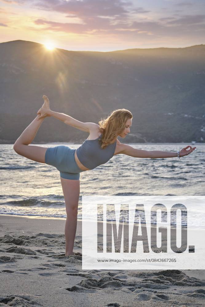 Eine Frau führt Natarajasana oder Dancer s Pose an einem Strand bei Sonnenuntergang auf. Die Sonne geht hinter den Bergen unter und schafft eine wunderschöne, friedliche Atmosphäre, während sie auf einem Bein balanciert A woman performs Natarajasana, or Dancer s Pose, on a beach during sunset. The sun sets behind mountains, creating a beautiful, peaceful atmosphere as she balances on one leg