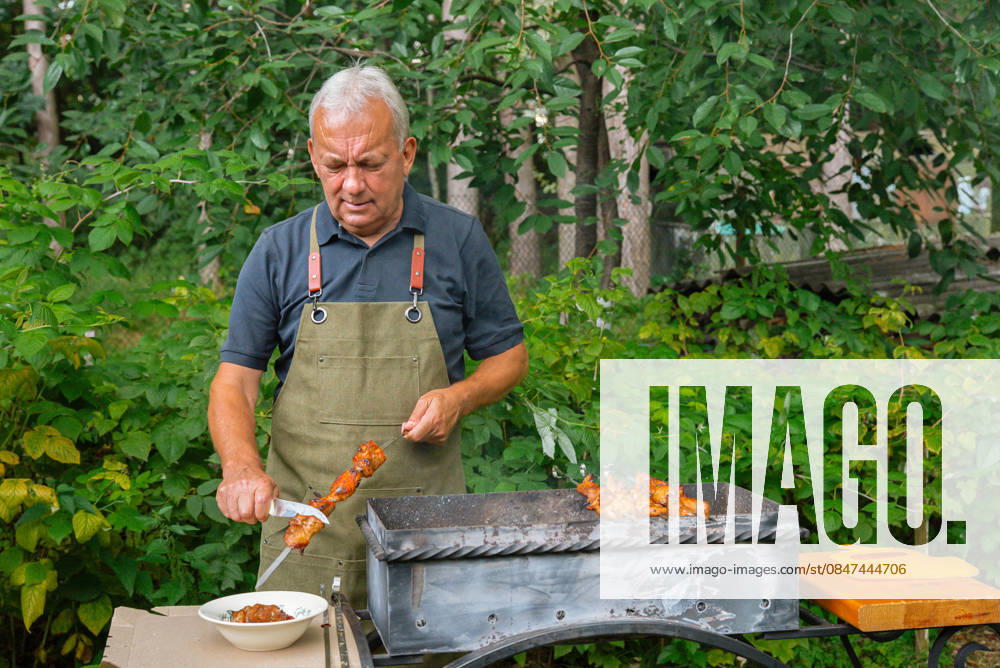 MR_ID32729_23803_AnatoliiIvanovich.jpg, MR_ID32729_23804_NilaAnatoliivna.jpg A man is preparing a barbecue in a lush garden, surrounded by greenery. He is focused on grilling and carving meat skewers, enjoying a relaxed outdoor cooking experience. Model Released Yanishevska_ID32729_1114335_033