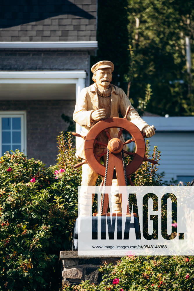 Tadoussac, Kanada ? 22. August 2025: Holzstatue eines Seemanns mit einem Schiffssteuerrad, ausgestellt in einem kleinen Garten im Dorf Tadoussac, umgeben von Blumen und traditionellen Häusern im Hintergrund. Tadoussac, Kanada ? 22. August 2025: Holzstatue eines Seemanns mit einem Schiffssteuerrad, ausgestellt in einem kleinen Garten im Dorf Tadoussac, umgeben von Blumen und traditionellen Häusern im Hintergrund. (License=RM)
