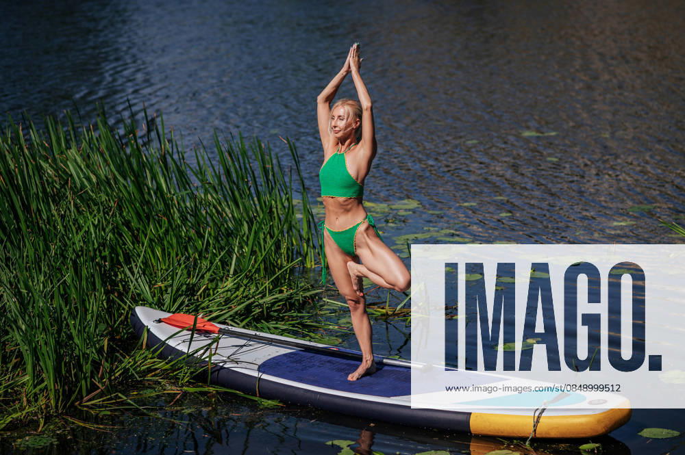 SUP yoga practice on a serene lake with paddleboard A woman practices SUP yoga on a paddleboard in a peaceful lake setting, surrounded by lush greenery. The tranquility of the scene highlights the harmony between nature and exercise. Model Released VilshanskyiBohdan_ID33046_1124666_012
