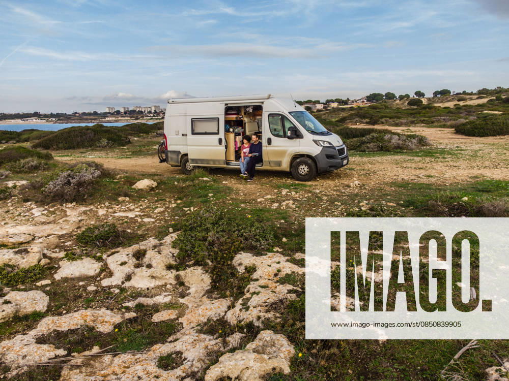 Camper sitzen in der Schiebetür ihres Campers auf einem unbefestigten Offroad Stellplatz an der Küste am Mittelmeer bei Tarent. Salento, Apulien, Italien. Ein Wohnmobil steht auf einem Schotterplatz am Ufer des Mittelmeers.