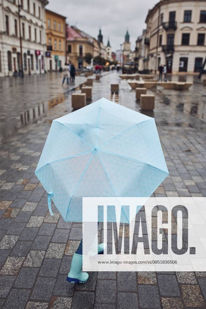 Rückansicht eines Kindes mit einem großen blauen Regenschirm, das an einem regnerischen, düsteren Herbsttag in einer Innenstadt spaziert Back view of child holding big blue umbrella walking in a downtown on rainy gloomy autumn day