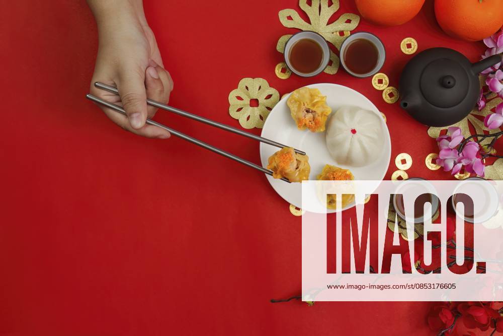 IMAGO Creative, Food, Dumplings Eine Hand, die mit Essstäbchen Knödel auf festlichem rotem Hintergrund aufhebt, symbolisiert die Traditionen und Freude des chinesischen Neujahrs A hand using chopsticks to pick up dumplings on a festive red background, symbolizing Chinese New Year traditions and joy