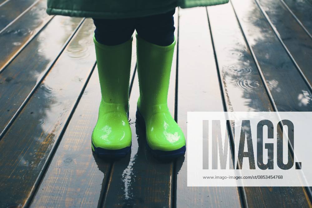 Nahaufnahme der Beine eines Kindes, das auf einer Veranda steht und grüne Gummistiefel und Regenmantel trägt. Regenstiefel in hellgrüner Farbe Closeup of legs of kid standing on a porch wearing green wellies and raincoat. Rain boots in bright green color