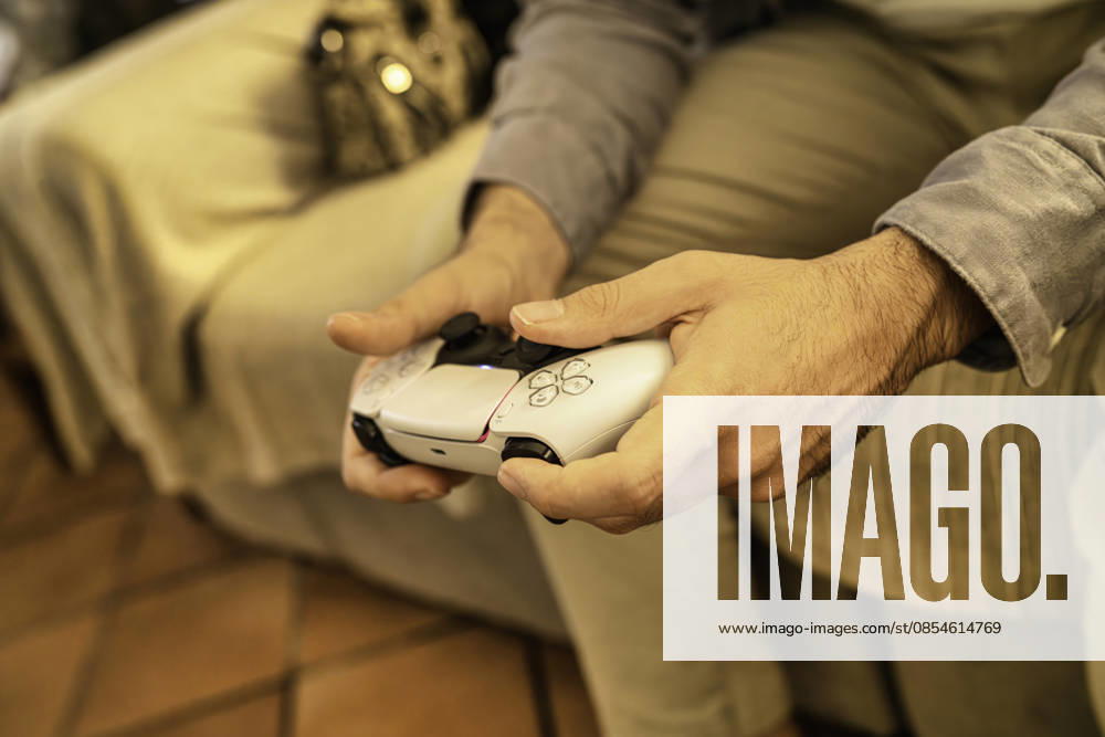 Milan, italy 28 october 2025: hands gripping a white ps5 dualsense controller while playing a video game at home for leisure and relaxed entertainment Person holding playstation 5 dualsense controller playing video game