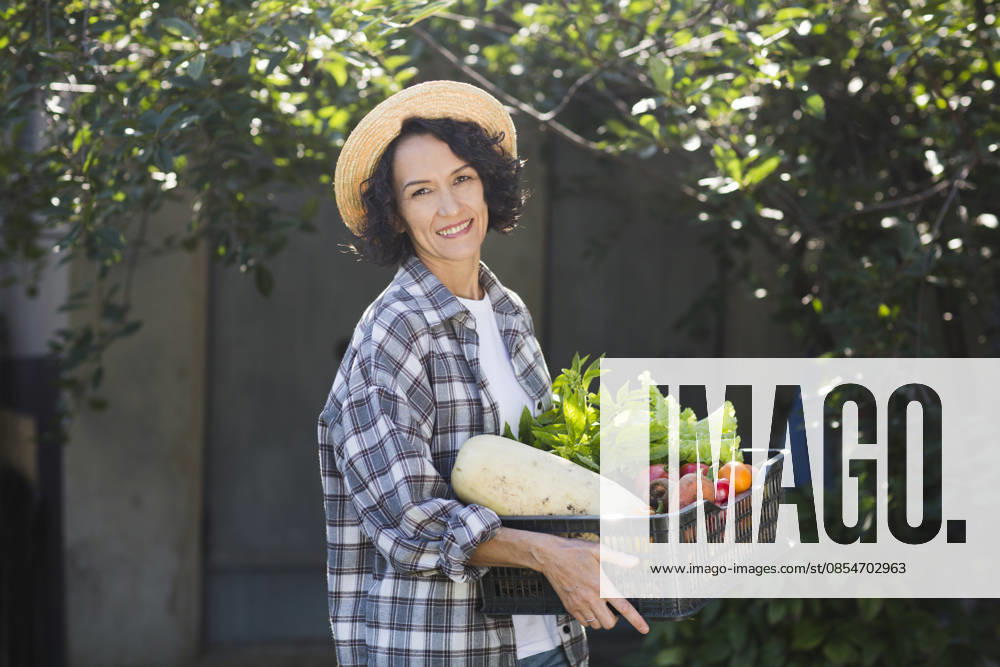 Eine fröhliche Frau mittleren Alters mit einem Strohhut hält eine Schale mit Gemüse in der Hand, das sie in ihrem Garten geerntet hat. Happy middle aged woman wearing a straw hat holds a box of vegetables picked in her garden.. (License=RF) ,model released, Symbolfoto