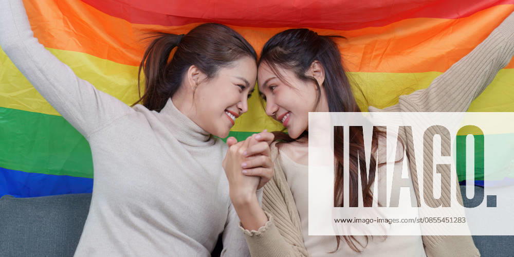 Ein asiatisches lesbisches Paar, das Händchen hält ? ein inniger Moment vor einer Regenbogenfahne An Asian lesbian couple shares a tender, intimate moment, holding hands and looking into each other s eyes, with a vibrant rainbow pride flag behind them. This image conveys love, connection, and LGBTQ pride. (License=RF) ,model released, Symbolfoto
