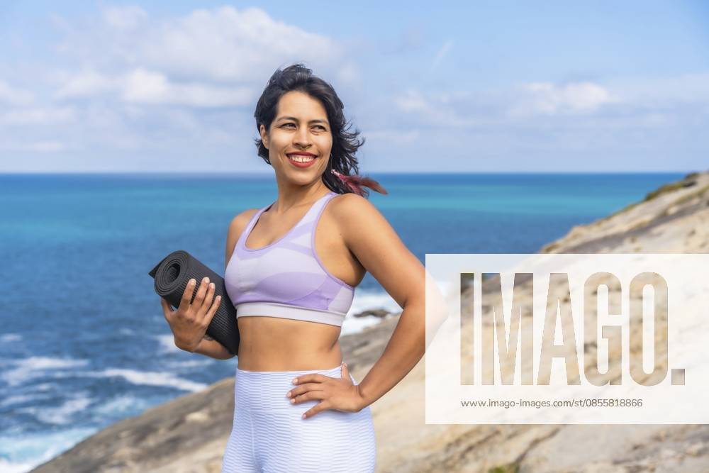 Frau lächelt, hält eine gerollte Yogamatte auf einer felsigen Klippe und blickt auf das strahlend blaue Meer, was für einen gesunden Lebensstil, Fitness und Wellness steht Woman smiling, holding a rolled yoga mat on a rocky cliff, looking towards the bright blue ocean, representing a healthy lifestyle, fitness, and wellness journey