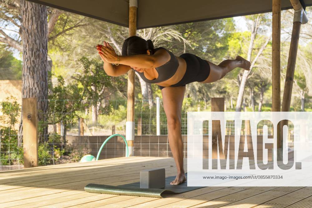 Die Frau hält das Gleichgewicht in einer Yoga Haltung auf einer Holzterrasse in einer ruhigen Umgebung im Freien aufrecht und fördert Wohlbefinden und Achtsamkeit durch körperliche Aktivität Woman maintains balance in a yoga posture on a wooden deck in a peaceful outdoor setting, promoting wellness and mindfulness through physical activity