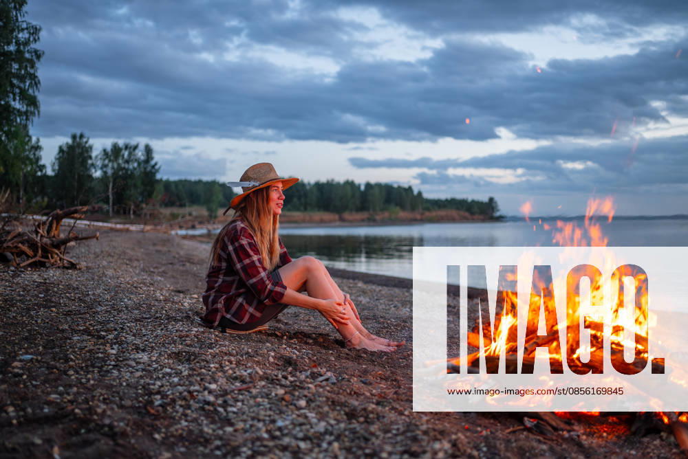 Eine Frau genießt einen ruhigen Abend am Lagerfeuer am Wasser; sie sitzt an einem Kiesstrand in der Nähe des Sees und genießt die Einsamkeit und Entspannung während eines Sommercampingausflugs Young woman sitting on a gravel lakeshore next to a crackling bonfire at dusk, finding warmth and relaxation while enjoying the calm nature and peaceful solitude of the outdoor evening (License=RF) ,model released, Symbolfoto
