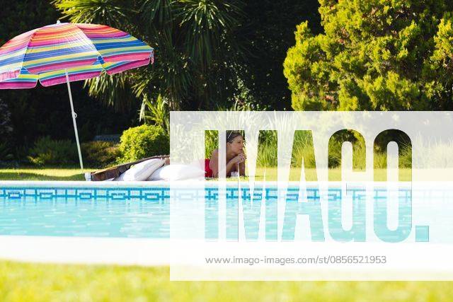 Adult woman lying on stomach at backyard pool edge wearing red swimsuit under striped umbrella, model released, property released, 19.03.2025 06:00:00, 12839384