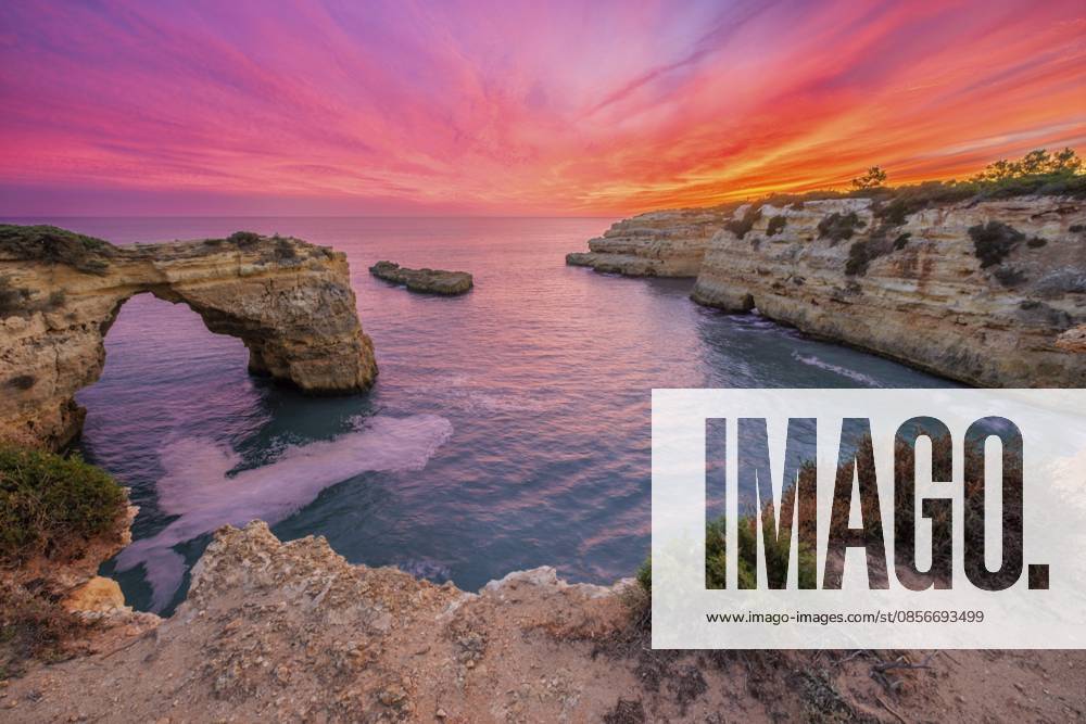 Dramatischer Blick auf den Sonnenuntergang von Arco de Albandeira am Praia de Albandeira an der Algarve, Portugal, mit rosafarbenem Himmel, Kalkstein Meeresbogen, Atlantikwasser und Dämmerungsfarben, die sich über der Bucht niederlassen Dramatic sunset view of Arco de Albandeira at Praia de Albandeira in Algarve, Portugal, with pink sky, limestone sea arch, Atlantic water, and twilight color settling over the cove