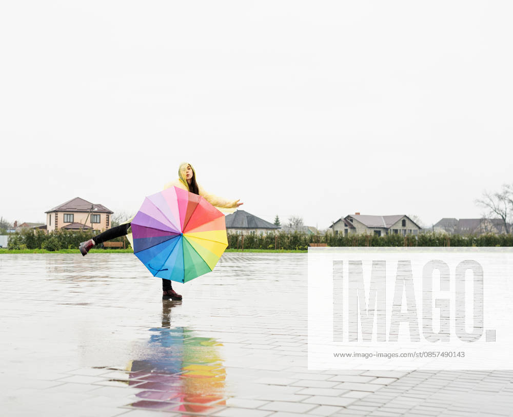 Eine schöne brünette Frau, die einen bunten Regenschirm hält und im Regen tanzt Beautiful brunette woman in yellow raincoat holding rainbow umbrella out in the rain (License=RF) ,model released, Symbolfoto