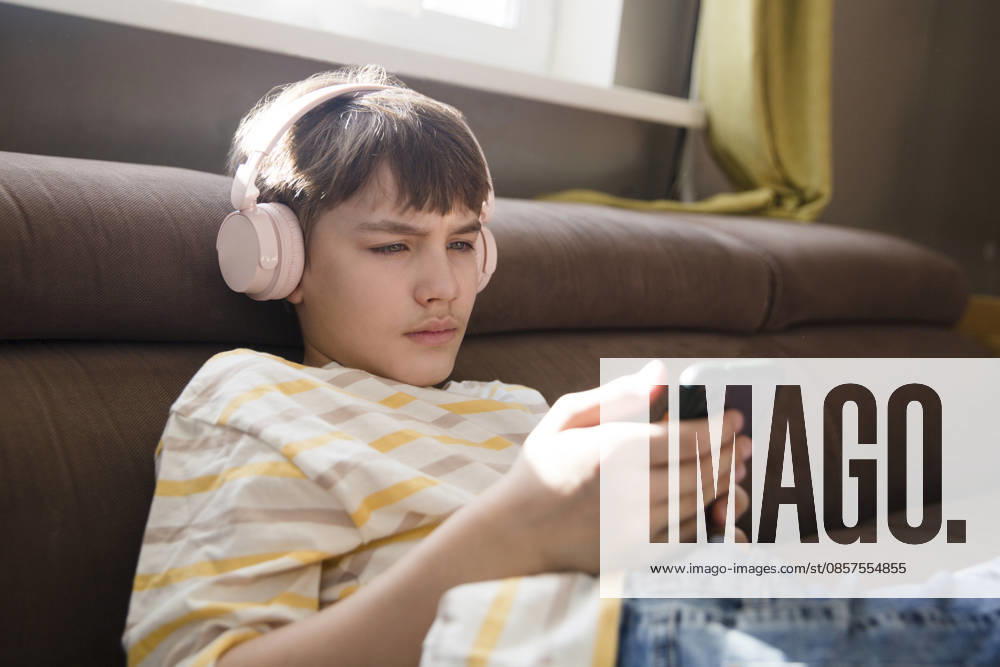 Ein Junge im Vorpubertätsalter sitzt zu Hause auf dem Sofa, hat Kopfhörer auf, hört Musik und schaut sich ein Video an. Seine Lieblingsmusik. Generation Z. preteen boy wearing headphones listens to music and watches a video sitting on the couch at home. favorite music. Generation Z. (License=RF) ,model released, Symbolfoto