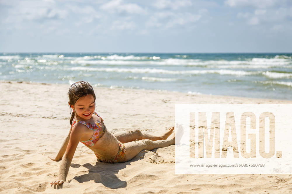 Ein süßes kleines Mädchen im Badeanzug sitzt am sandigen Strand, mit Sand bedeckt, während hinter ihr die Meereswellen brechen ? ein Bild, das die Freude an den Sommerferien und unbeschwerte Kindheitserinnerungen verkörpert Ein süßes kleines Mädchen im Badeanzug sitzt am sandigen Strand, mit Sand bedeckt, während hinter ihr die Meereswellen brechen ? ein Bild, das die Freude an den Sommerferien und unbeschwerte Kindheitserinnerungen verkörpert (License=RF) ,model released, Symbolfoto