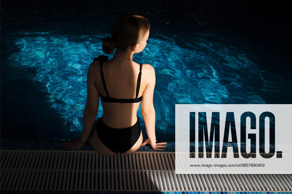 Eine weibliche Figur in einem schwarzen Badeanzug sitzt am Beckenrand, von Sonnenlicht beleuchtet, während das schimmernde Wasser das Licht in einer ruhigen Hallenbadumgebung reflektiert Woman in black swimsuit sits on the edge of a pool, sunlight creates reflections on the water surface, creating a tranquil atmosphere in the indoor setting (License=RF) ,model released, Symbolfoto