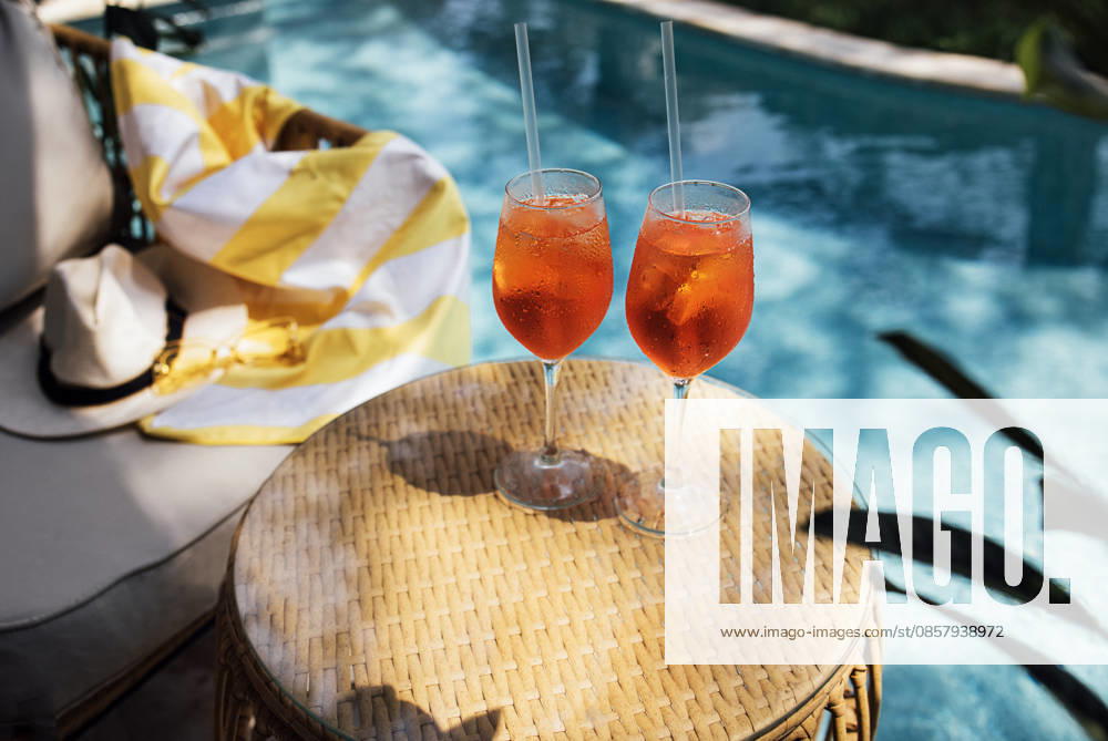 Zwei erfrischende Aperol-Spritz-Cocktails mit Orangenscheiben und Strohhalmen auf einem geflochtenen Tisch am Pool, im Hintergrund ein bunter Handtuch über einem Liegestuhl Two aperol spritz cocktails with orange slices and straws sit on a woven table next to a swimming pool, with a colorful towel on a lounge chair nearby (License=RF)