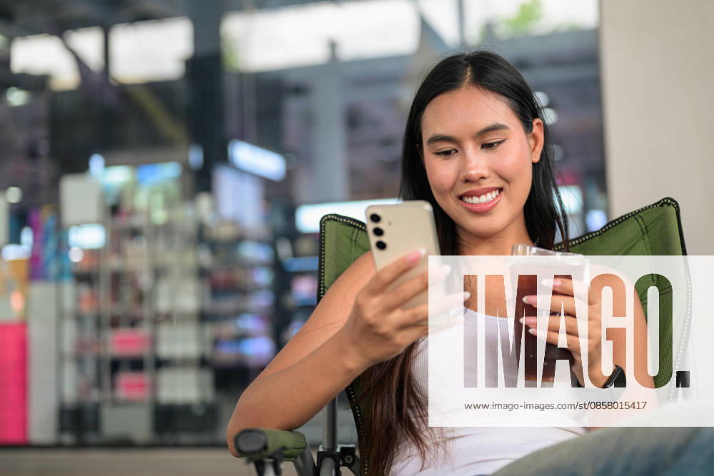 IMAGO Creative, Food, Trend, Iced Coffee Eine lächelnde Thailänderin, die im Freien ihr Smartphone benutzt, während sie einen Eiskaffee in der Hand hält. Horizontal medium shot of a happy young Isan Thai female customer looking down and smiling while operating her smartphone held in both hands. Seated relaxed in a green folding camping chair outdoors, she concurrently holds a clear plastic takeaway cup of iced coffee or iced tea, featured in a wide composition with excellent copy space on the left side. (License=RF) ,model released, Symbolfoto