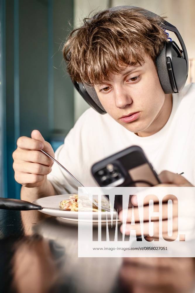 Ein Teenager mit Kopfhörern, der beim Mittagessen zu Hause auf sein Smartphone schaut Focused teenage boy wearing headphones uses his smartphone while having a meal at the kitchen table, distracted by digital content. (License=RF) ,model released, Symbolfoto