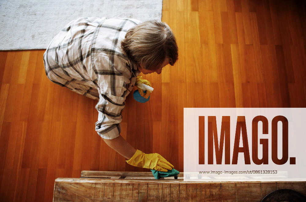 ClelaninnnngDuusttttttty.jpg An elderly woman in casual clothes washes a wooden coffee table in the living room. A mature lady is cleaning the house. A pensioner in rubber gloves cleans the countertop. Top view. A cozy interior (License=RF) ,model released, Symbolfoto