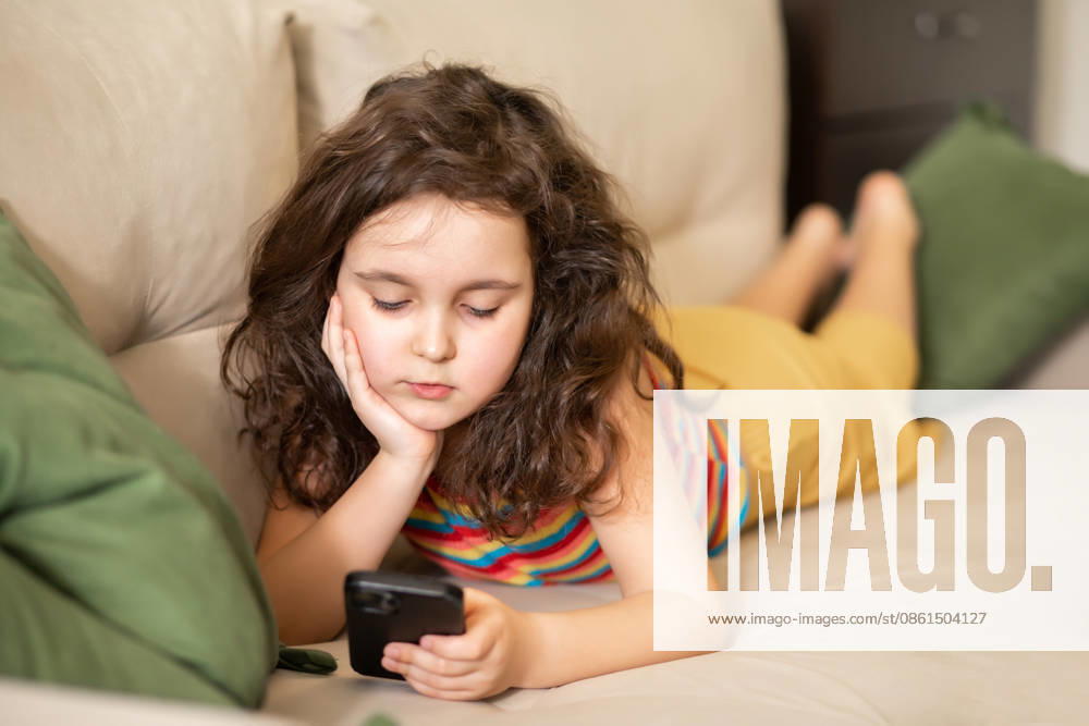 Ein süßes kleines Mädchen, das mit seinem Smartphone spielt, liegt zu Hause auf dem Sofa im Wohnzimmer Bored and sad cute little girl using smart phone lies on sofa in living room at home. (License=RF) ,model released, Symbolfoto
