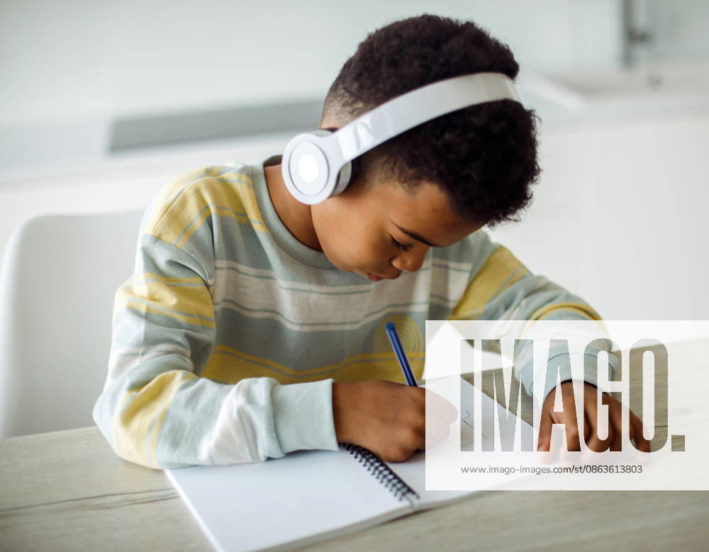 Ein Junge gemischter Herkunft schreibt in ein Notizbuch und hört über Kopfhörer Musik A mixed race boy writes in a notebook and listens to music on headphones at home. (License=RF) ,model released, Symbolfoto