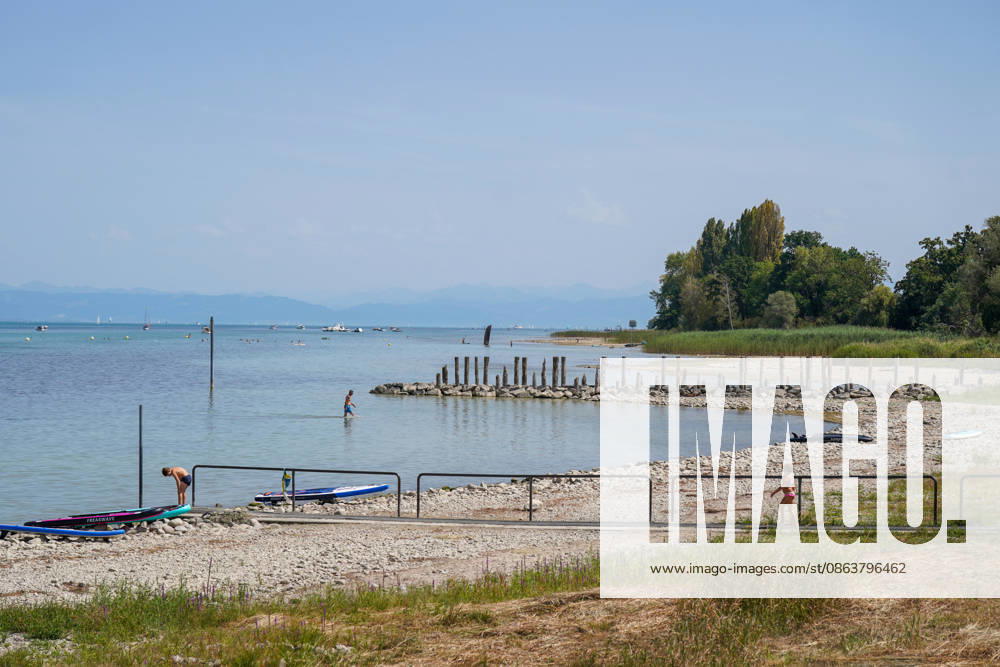 Schweiz, Bodensee: Niedriger Wasserstand legt breite Kies- und Uferflächen frei. Badegäste stehen im flachen Wasser, SUP-Boards liegen am Ufer; im Hintergrund Boote und Alpenpanorama. Symbolbild für Bodensee, Wasserstand, Niedrigwasser, Trockenheit, Hitze, Hitzewelle, Sommer, Klimawandel, Gewässer, Ufer, Kiesstrand, Baden, Abkühlung, Freizeit, Wassersport, Natur, Umwelt, Schweiz.