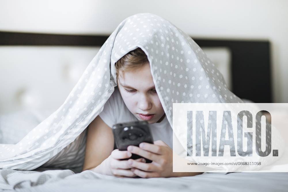Süßer kleiner Junge mit Smartphone liegt auf dem Bett unter dem Cover zu Hause Cute little boy with smartphone lying on bed under cover at home