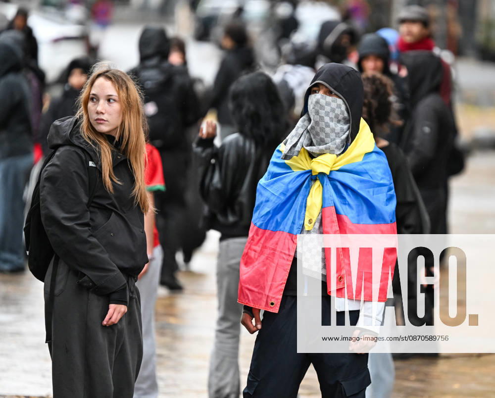 A demonstrator wearing a Venezuelan flag as a cape stands among a crowd during student protests in central Brussels, Belgium, on 24 October 2026. The man wears a black hood and a face covering, while a young woman stands beside him. Other demonstrators gather in the background during protests that led to repeated police interventions across the city centre.