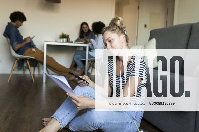 IMAGO Creative, Lifestyle, Leben in einer Wohngemeinschaft, WG Young woman sitting on floor with cell phone and papers and friends in background model released Symbolfoto property released VABF01780