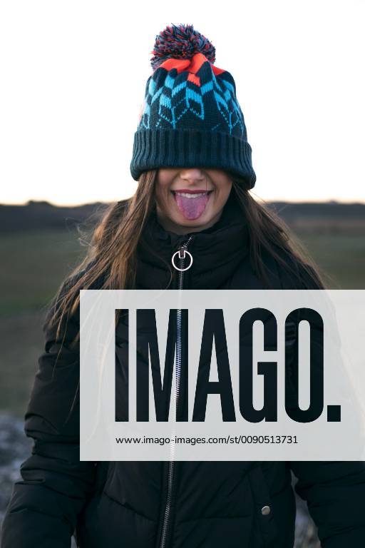 Teenage girl wearing multicolored bobble and black down jacket sticking out tongue model released Symbolfoto ACPF00377
