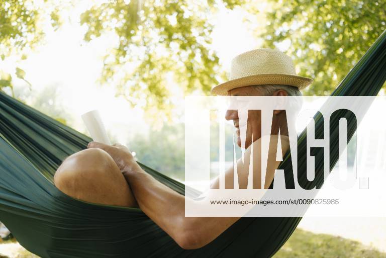 Senior man wearing straw hat relaxing in hammock at lakeshore reading book model released Symbolfoto property released GUSF01788