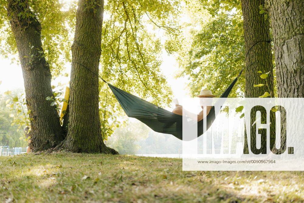 Senior man wearing straw hat relaxing in hammock at lakeshore reading book model released Symbolfoto property released GUSF01786