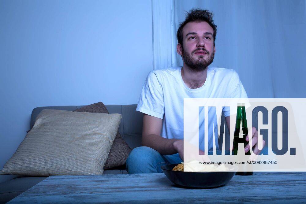 A young man watching with passion TV at nighttime with potato chips and beer at home in the living room.,model released, Symbolfoto